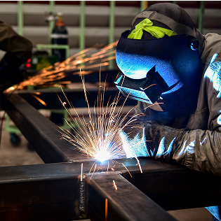 Welder wearing mask welding metal frame, sparks flying in industrial setting.