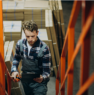 Warehouse worker with clipboard climbs stairs near stacked boxes.